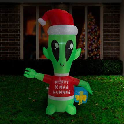 Inflatable green alien with Santa hat holding a blue gift in front of Christmas decor