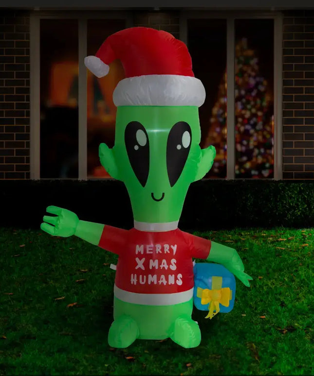 Inflatable green alien with Santa hat holding a blue gift in front of Christmas decor