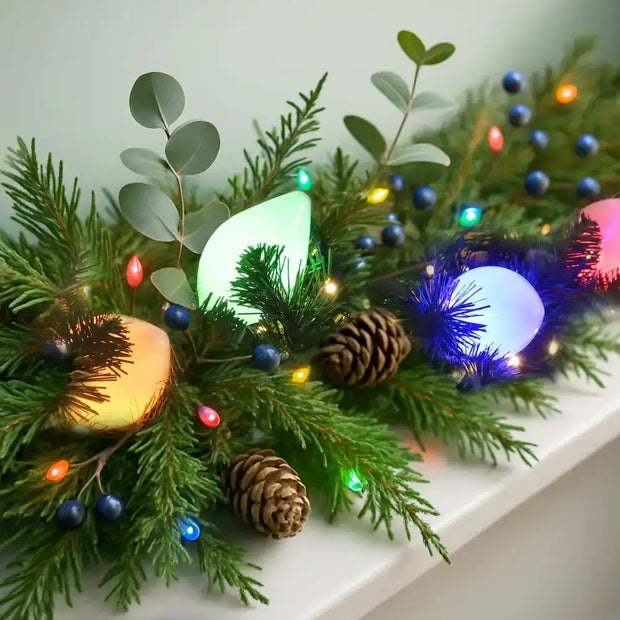 A festive garland featuring glowing, colorful led lights nestled among green pine branches and blue berries.